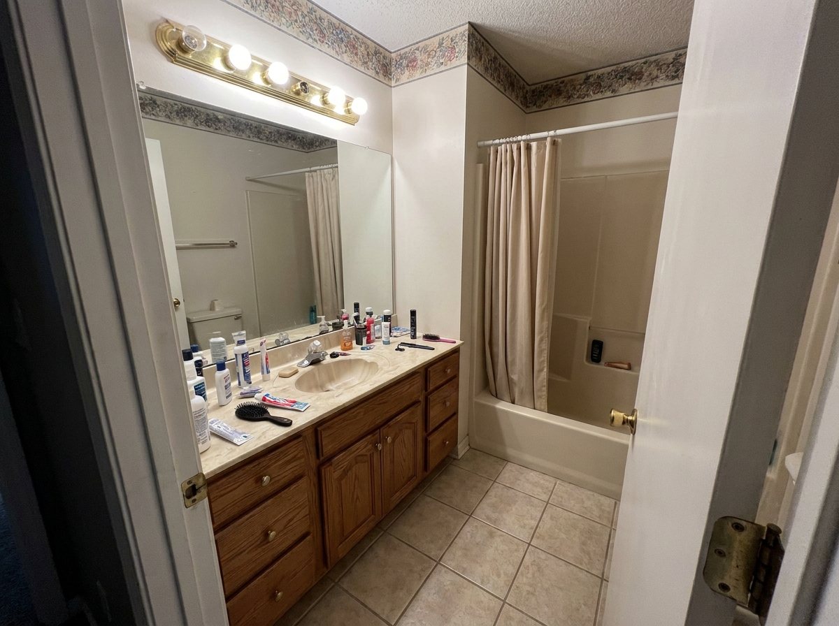 A dated small bathroom with a floral wallpaper border, oak vanity, brass bulb light bar and beige tub surround